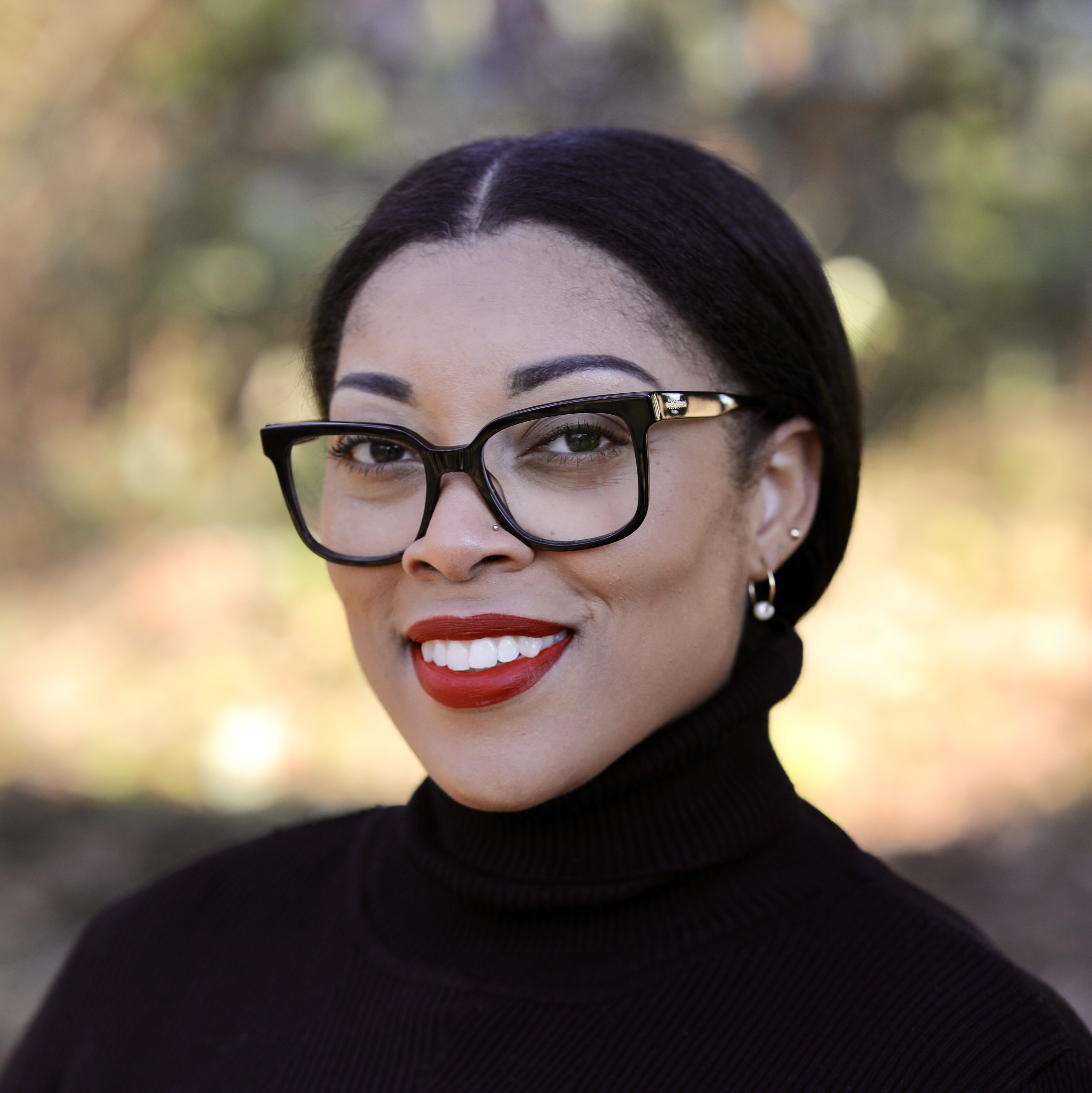 A woman with glasses and red lipstick smiles, wearing a black turtleneck, set against a blurred outdoor background.
