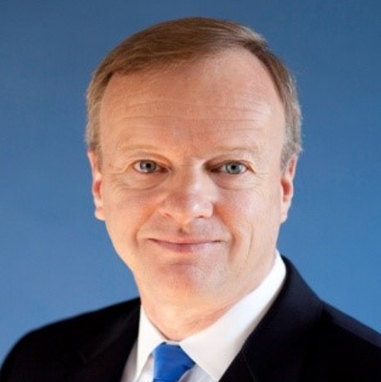 A professional headshot of a man with short, light brown hair, smiling in a dark suit and blue tie against a blue backdrop.