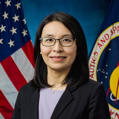A woman with black hair and glasses smiles, wearing a black blazer over a purple blouse, against NASA flags and a blue background.