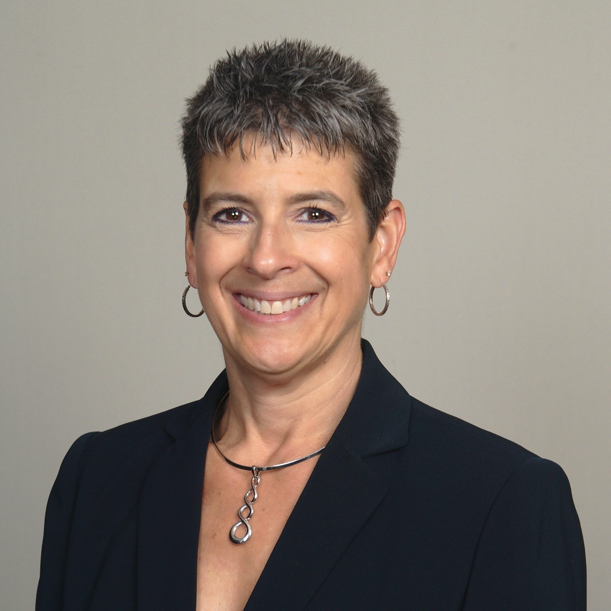 Smiling woman with short, spiky gray hair, wearing a dark blazer over a pink top, set against a neutral gray background.