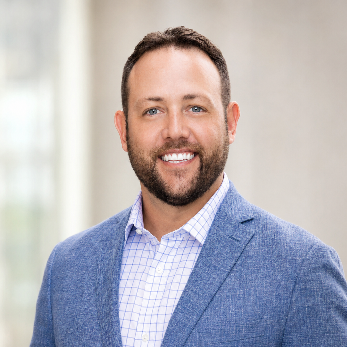 Smiling man with a beard in a fitted blue blazer over a plaid shirt, posed against a softly blurred light background.