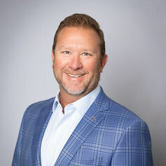 Smiling man in a blue plaid blazer and white shirt, set against a light gray background.
