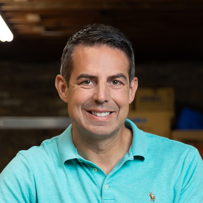 Man smiling against a blurred background, wearing a teal polo shirt with a small logo, short dark hair, and a friendly expression.