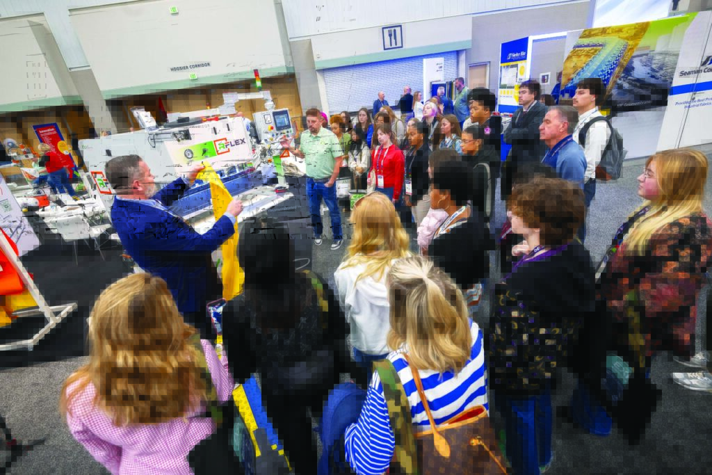 A diverse group of attendees gathers around the Miller Weldmaster booth at Advanced Textiles Expo, showcasing the company's machinery. Many textiles program college students are paying close attention.