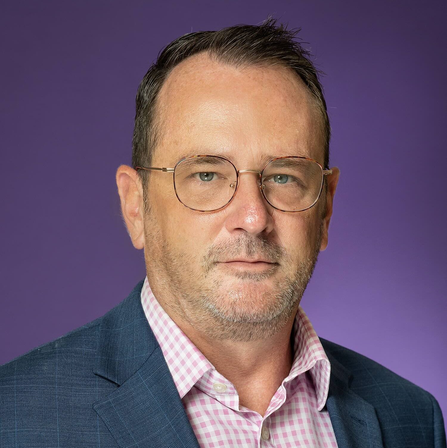 A man with glasses and short hair in a blue blazer over a pink checkered shirt, set against a purple background.