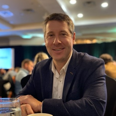 Smiling man in a blazer at a dining table, with a glass of water and a bowl in front; softly lit background with event attendees.