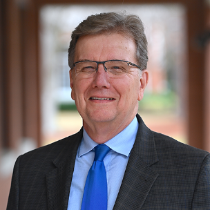A middle-aged man with glasses smiles, wearing a blue tie and a dark plaid suit, set against a blurred outdoor background.