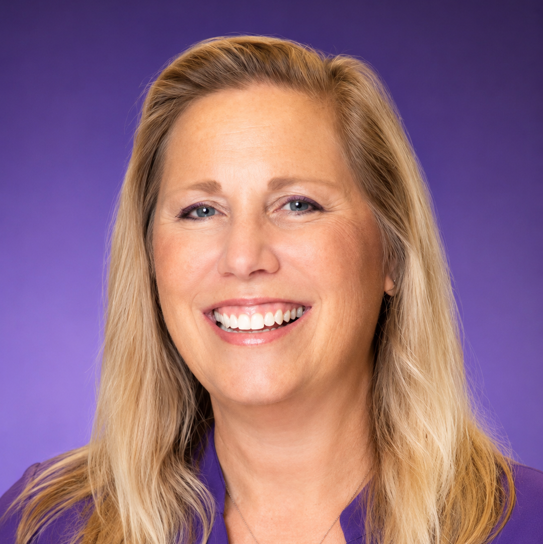 Smiling woman with long blonde hair, wearing a purple top, against a solid purple background.