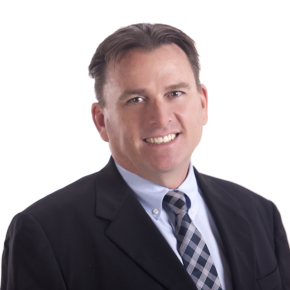 Man in a black suit jacket with a light blue dress shirt and patterned tie, smiling against a white background.