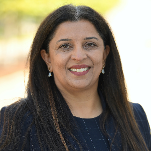 Portrait of a woman with long dark hair, wearing a navy blazer, smiling against a softly blurred outdoor background.