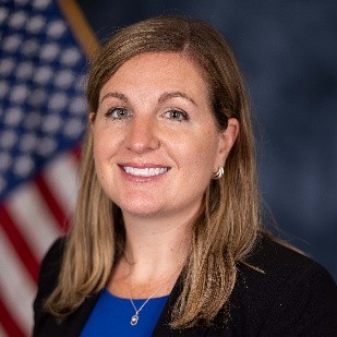 Headshot of a woman with shoulder-length, light brown hair, wearing a black blazer over a blue shirt, smiling against a blurred American flag background.